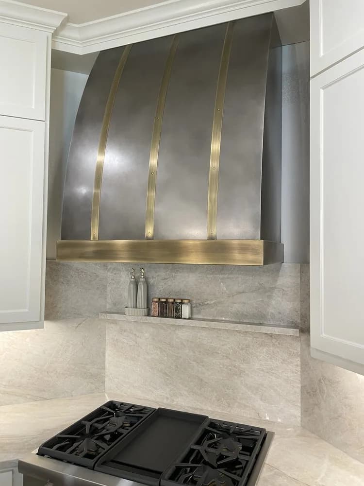 Antiqued stainless steel range hood with brass straps above a cooktop, set between white cabinetry and a marble surround.