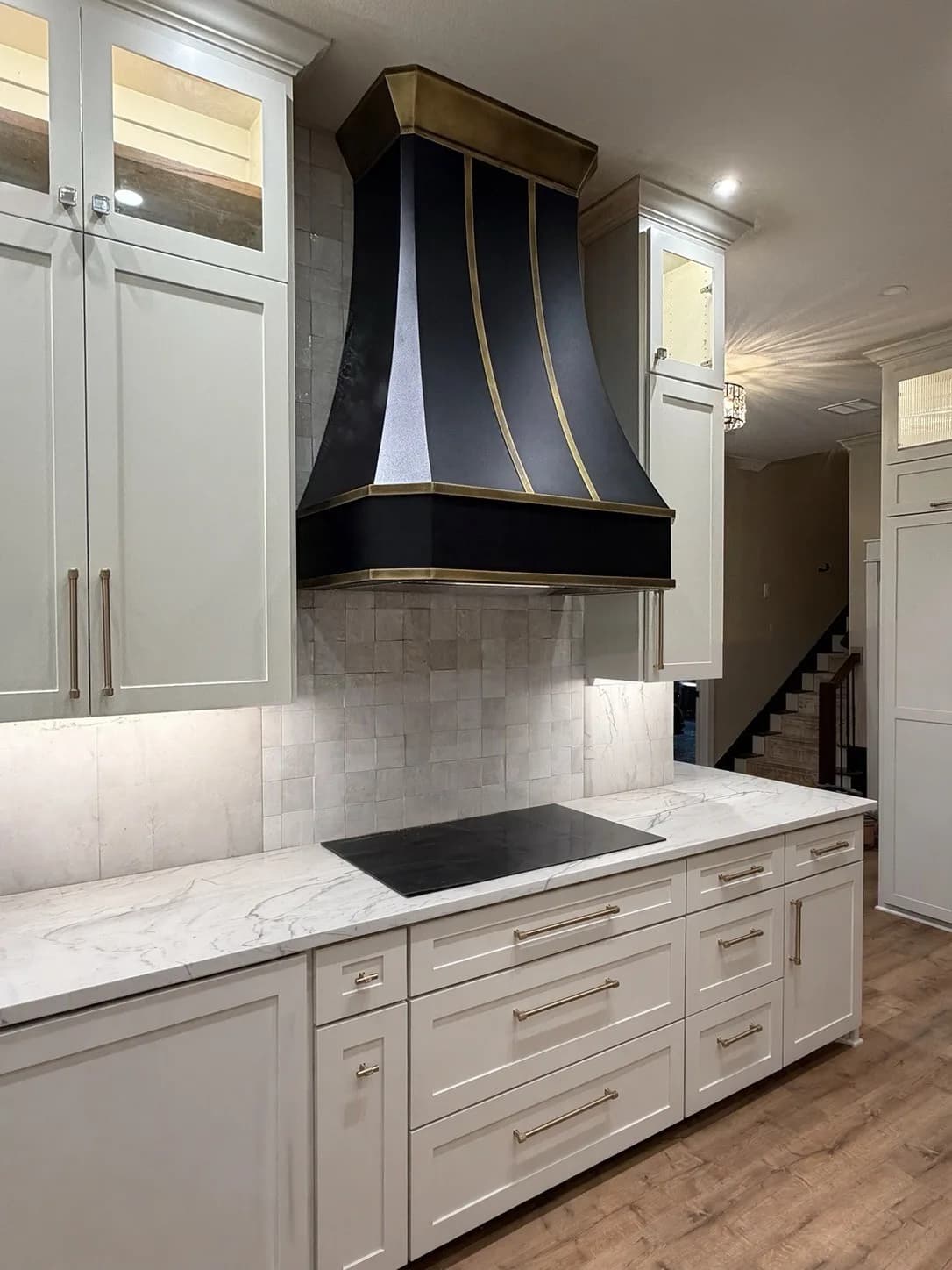 Custom black range hood with brass straps and banding over a zellige tile backsplash, in a white kitchen with marble countertops.