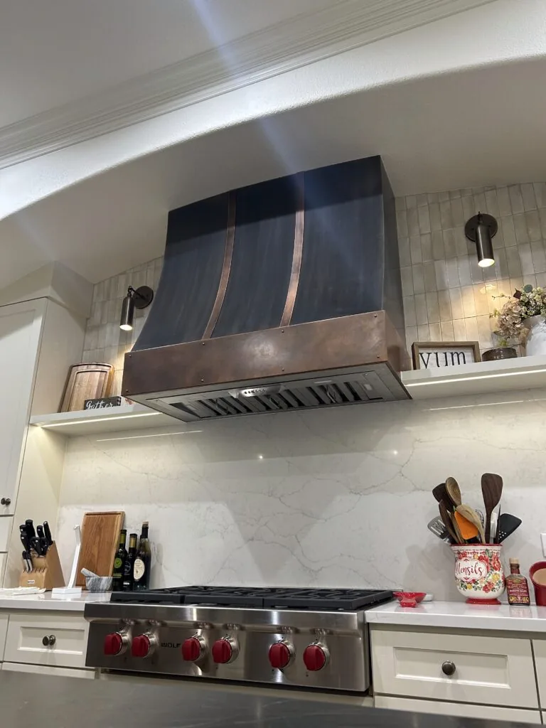 Copper range hood with a dark band above a wide professional gas range, flanked by open shelving and a marble backsplash.