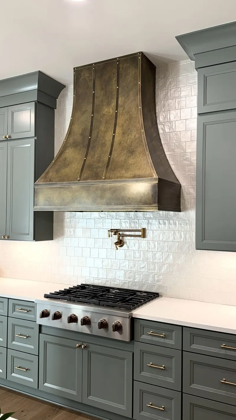 Hand-patinated brass range hood above a gas range, between sage green cabinets and a white tile backsplash