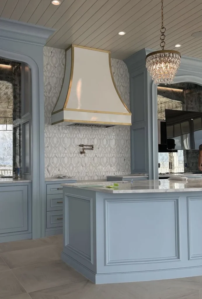 Pale powder coated range hood in a light kitchen with a painted island, coffered ceiling and a glass pendant.