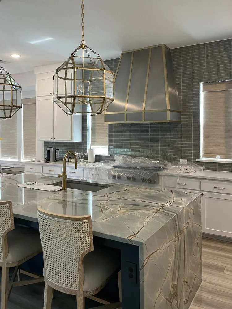 Wall mounted custom metal range hood in an open kitchen with a large island, lantern pendants and tiled walls.